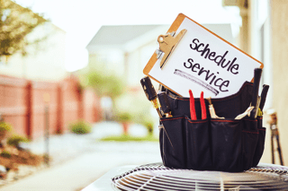 Why Schedule Air Conditioning Maintenance? Toolbox sitting on top of an air conditioning unit with a clip board that reads, "Schedule Service,” on it.