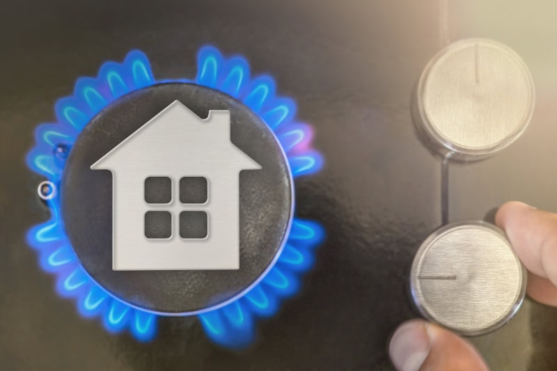 A hand adjusting the dial of a lit gas stove burner overlaid with a small house icon.